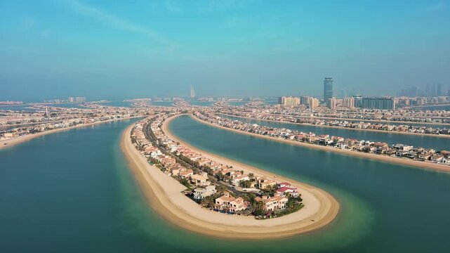 Aerial high-angle panoramic video of Palm Jumeirah frond ends and distant Dubai Marina skyline. Luxury waterfront villas and calm Arabian Gulf water on sunny day.
