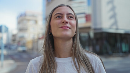 Young woman looking up on city street, face and bare neck visible, wearing white t shirt and gold hoop earring while standing near buildings  serenity. © Krakenimages.com