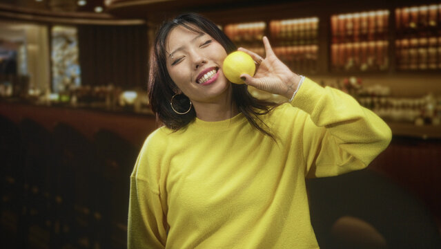 Young woman holding lemon near cheek, smiling with closed eyes and raised hand in studio bar set; playful curiosity.