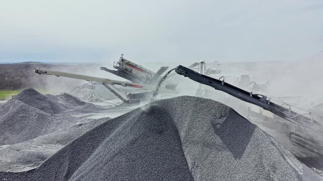 Heavy machinery works in a granite quarry to crush rock into gravel while dust fills the air and piles are formed.