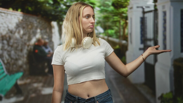 Woman with headphones showing palm up gesture and exposed midriff on narrow street beside white wall and green bench; indifference waiting.
