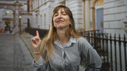 Woman points finger upward on city street beside iron railing, wearing striped shirt and hoop earrings with visible hand gesture and broad smile  confidence advice. © Krakenimages.com