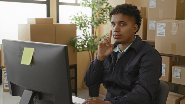 Young hispanic man at computer surrounded by parcel boxes, typing and finger to lips for silence in shipping building; concentration.