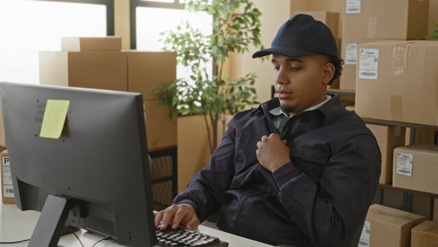 Man typing at computer in a packing building with stacked boxes and parcels, right hand raised in a wave; friendly productivity.