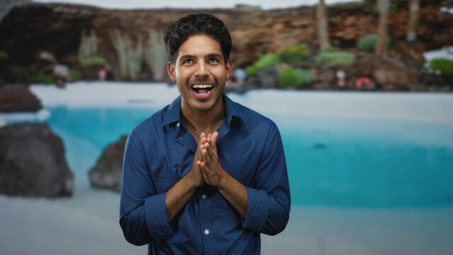 Man smiling with hands clasped, blue shirt, standing before a tropical pool backdrop in studio; calm confident joy.