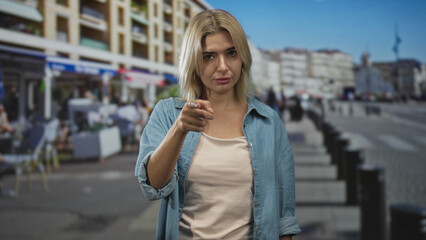 Fototapeta premium Woman pointing finger at camera on a crowded street wearing denim shirt and beige tank top; accusation.