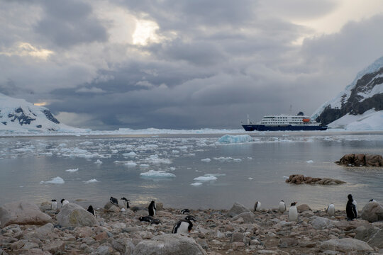 MV Hondius Expedition cruise ship navigating through icy waters in Antarctica, exploring melting glaciers and snow-capped mountains under a dramatic cloudy sky with sun breaking through