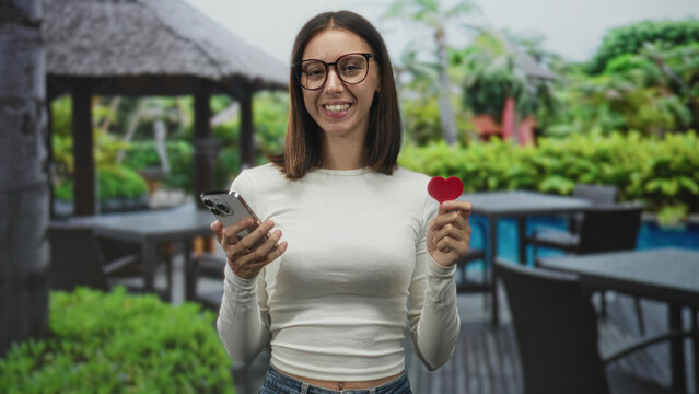 Young hispanic woman holds smartphone in left hand and small red heart in right hand, smiling and looking at phone, standing by pool tables and gazebo on building patio; digital romance joy.