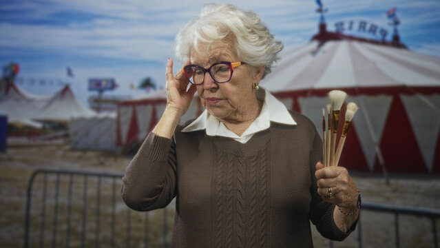 Elderly woman holding paintbrushes touches eyeglasses on street in front of circus tent, slight frown and thoughtful gaze; nostalgia calm.