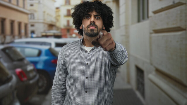 Man points finger at camera on street, curly hair and beard visible; confrontation anger accusation.