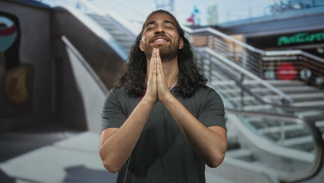 Man pressing bare hands together praying by an escalator inside a shopping mall building, eyes closed and head tilted back; gratitude prayer.