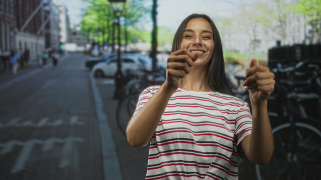 Young woman hands mimes steering motion on city street while smiling and reaching forward; joy playfulness.