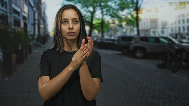 Woman claps hands with arms on hips and rolls eyes on a cobblestone street wearing a black t shirt; annoyance impatience sarcasm.