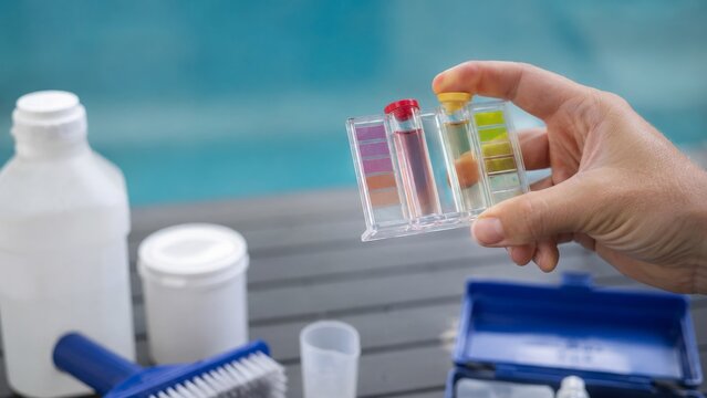 Hand holds a clear pool water test comparator with colored reagent vials and chart strips near bottles and a blue case beside swimming pool.