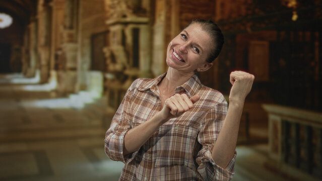 Woman smiling and holding thumb pointing to the side in an ornate church building corridor with visible hands and plaid shirt; joy.