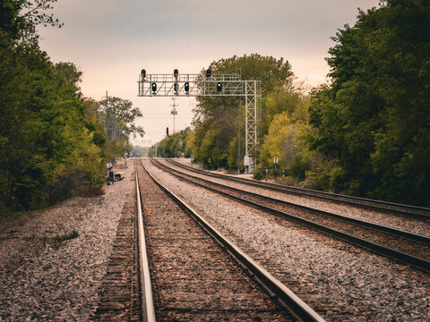 Double railway tracks receding into the distance under a signal bridge and green trees on a cloudy day in the Chicago suburbs