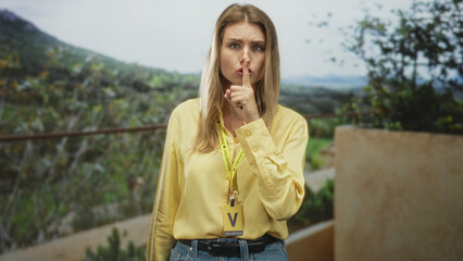 Naklejka premium Blonde woman in yellow shirt and jeans wearing volunteer badge and visitor card, finger to lips for silence in building overlooking vineyard hillside; serious warning.