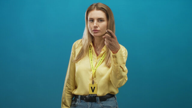Blonde woman points with hand showing volunteer badge and yellow lanyard idcard while facing camera in studio; assertive guidance.