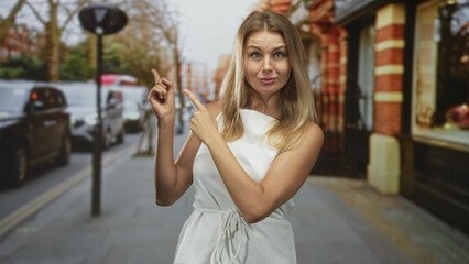 Fototapeta premium Woman points finger to left on city street in front of brick building wearing white dress; playful confidence.