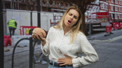 Fototapeta premium Woman holding a model brain in one hand, clutching stomach with the other, grimacing on street near construction barriers and scaffolding; mental health discomfort.