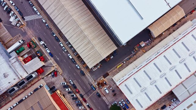 A busy street in an industrial zone with cars parked along the sides. There are warehouses on both sides under clear skies. The setting is active and bustling.