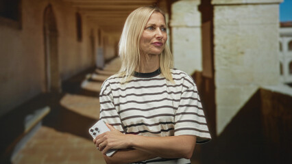 Fototapeta premium Woman holding smartphone, arms crossed and looking up in building corridor; contemplation quiet serenity.