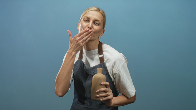 Woman artisan holds a handmade clay bottle and blows a kiss with hand to lips in studio while smiling and wearing apron; craft pride.