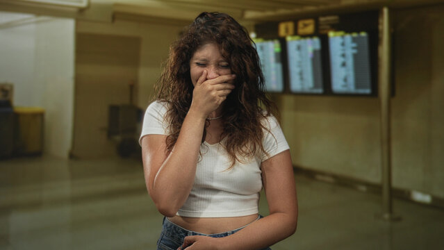 Woman covering mouth with hand, eyes squinted and torso slightly bent in airport terminal by flight information screens, wearing white crop top and jeans; embarrassment.