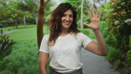 Naklejka premium Woman waving open hand on park path while smiling with palm visible and forearm shown; friendly greeting happy.