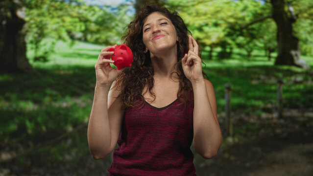 Young hispanic woman smiling while holding a red piggy bank and crossing fingers in a green park forest; saving hope.