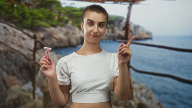 Woman with short buzz haircut holding a pink disposable razor near face with hand on chin, wearing white cropped top in studio; grooming contemplation.