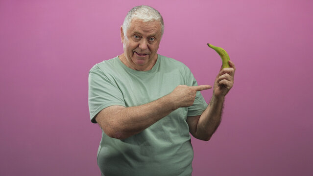 Senior man points finger to banana while holding it with other hand in studio pink backdrop, smiling and engaging directly with camera; playful.