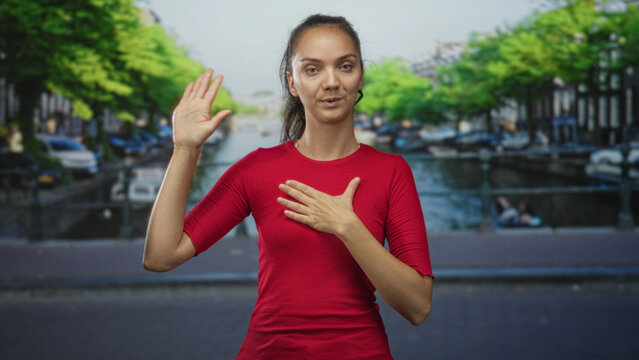 Woman with raised right hand and left hand on chest on street making oath pledge gesture in front of canal and bridge; sincerity apology.