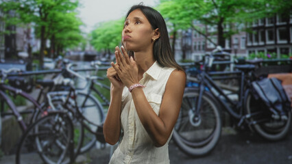 Fototapeta premium Thai woman clasping hands near chest beside parked bicycles on a canal street, looking upward with pleading gaze; hopeful prayer.