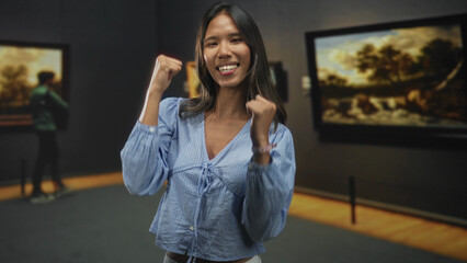 Fototapeta premium Thai woman with fists raised, smiling and dancing in a museum building among paintings; joy celebration.