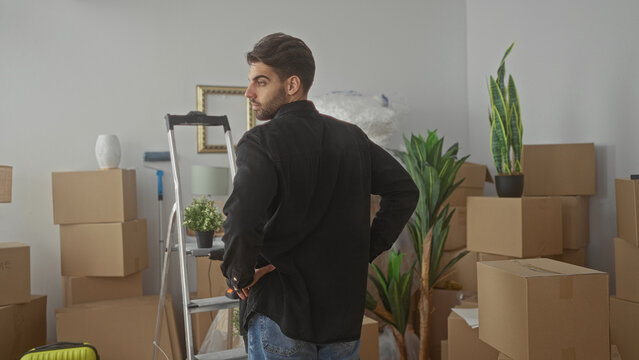 Man with beard standing hands on hips amid stacked cardboard boxes, ladder and potted plants in a building interior prepared for moving; planning uncertainty.