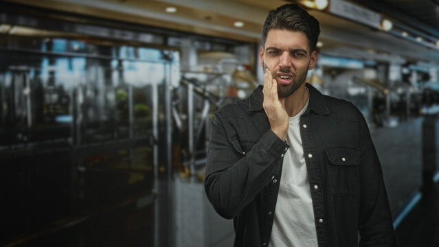Hispanic man pressing cheek with hand for toothache at airport check in queue, visible wince and closed eye; toothache discomfort travel.