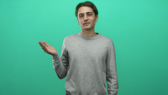 Man standing in studio wearing glasses and grey sweater displays palm up hand gesture with open empty hand, slight smile and relaxed posture; relaxed offering.