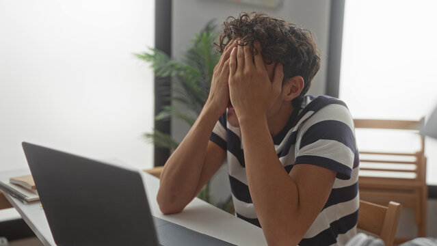 Man in striped shirt covering face with hands at desk with laptop and notebooks in building; stress exhaustion.