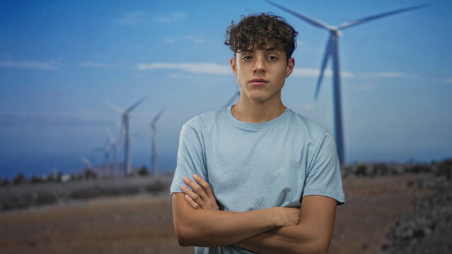 Teen boy in light blue tshirt with crossed arms and visible forearms standing by wind turbines on a street under a blue sky; quiet determination.