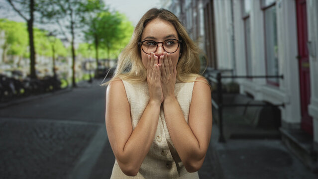 Woman with hands covering mouth and glasses on city street beside row of buildings and parked bicycles, light summer outfit; surprise curiosity.