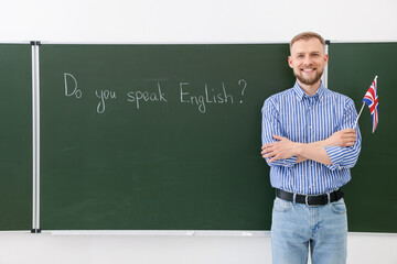 Obraz premium English teacher holding flag of United Kingdom at green chalkboard with phrase Do You Speak English? in classroom