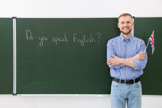 English teacher holding flag of United Kingdom at green chalkboard with phrase Do You Speak English? in classroom