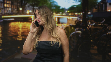 Fototapeta premium Middle aged woman holding phone to ear, eyes closed, wearing strapless black dress on street by lit canal and parked bicycles at night; frustration fatigue.