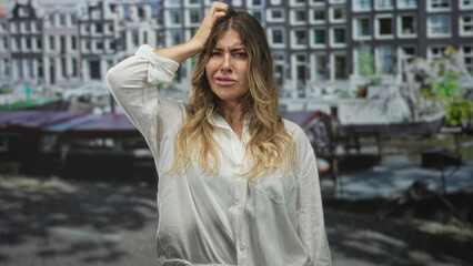 Fototapeta premium Woman holding head with hand and puzzled frown in front of canal building facade, loose white shirt visible, full body visible; frustration.