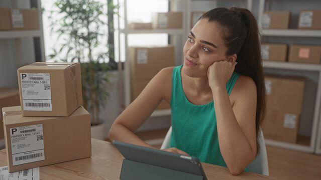 Woman with a stack of cardboard packages resting chin on hand beside tablet in building; contemplation.