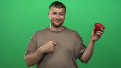 Fototapeta premium Young caucasian man holding a red apple with right hand and thumb pointing to chest in green studio; confidence healthy choice.