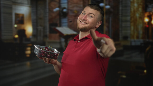 Man holding cherries points finger to camera in a church building, smiling and offering fruit; playful invitation.