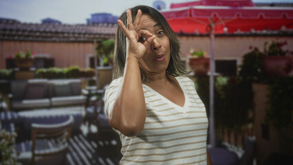 Fototapeta premium Hispanic woman shows hand making ok sign over eye on a building terrace with patio furniture and red umbrella; playful.