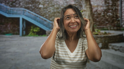 Fototapeta premium Woman adjusts glasses with hands, smiling and tucking hair while standing by a building courtyard; joy warmth.